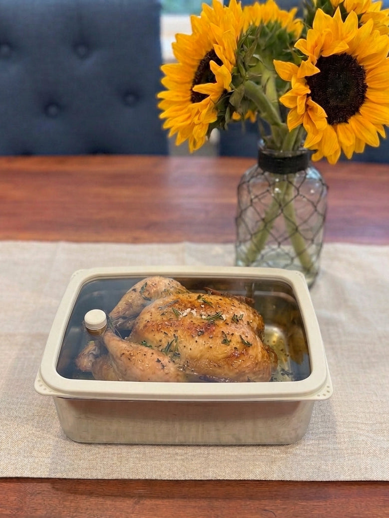A rectangular beige ceramic or stone baking dish with a clear glass lid, containing a roasted whole chicken, set on a beige placemat on a wooden table. A glass vase with sunflowers is in the background.
